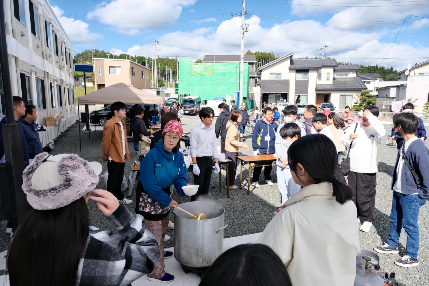 晴天の住宅街で開かれた交流イベントの場で、地域の女性が大鍋で温かい汁物をよそい、高校生や住民たちが周囲に集まり、賑わいと温もりに包まれた炊き出しの様子の写真