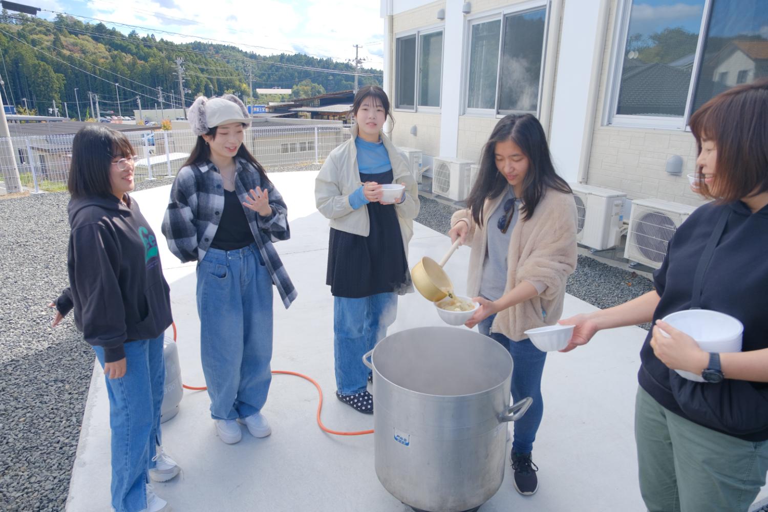 屋外で大きな鍋から女性がスープをお椀に注ぎ、他の女性たちが楽しそうに待っている地域の交流イベントの写真