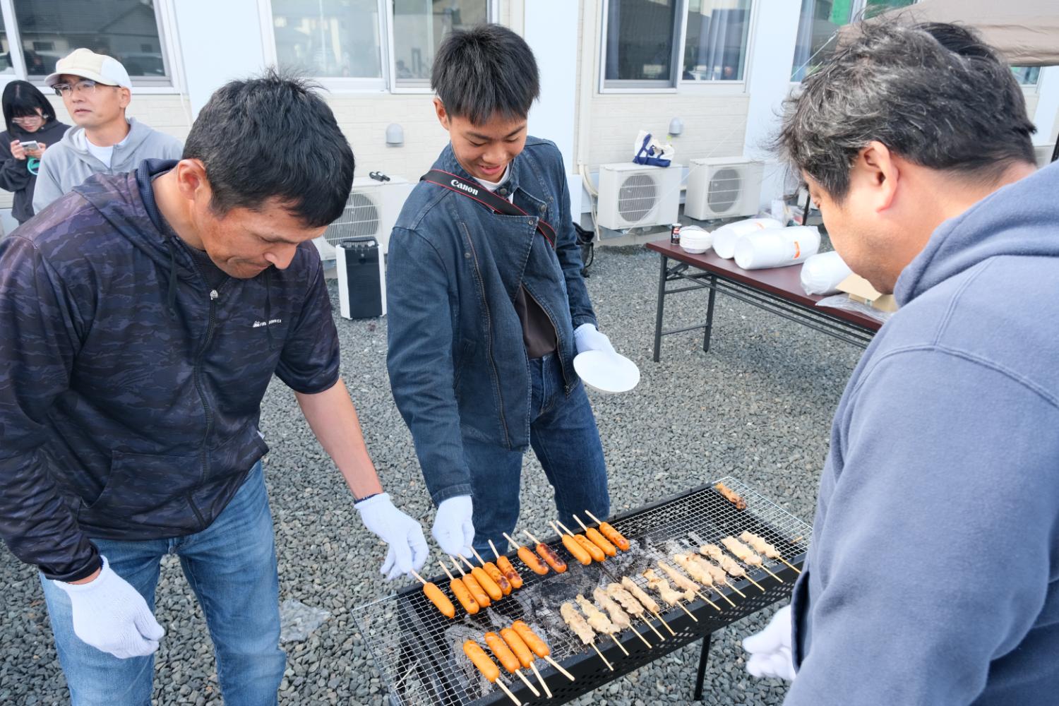 屋外の砂利敷きの場所で、白い手袋をはめた3人の男性が串に刺した焼き鳥やソーセージを網焼きグリルで焼いている様子の写真