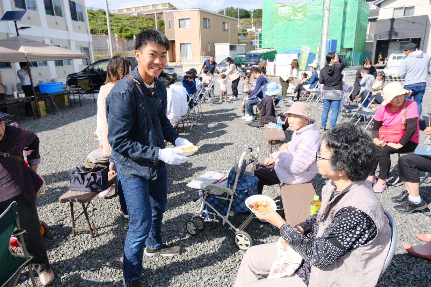 野外イベントで男性が白い手袋をはめて高齢者に料理を手渡し、多くの参加者が椅子に座って交流している地域のふれあい交流会の写真
