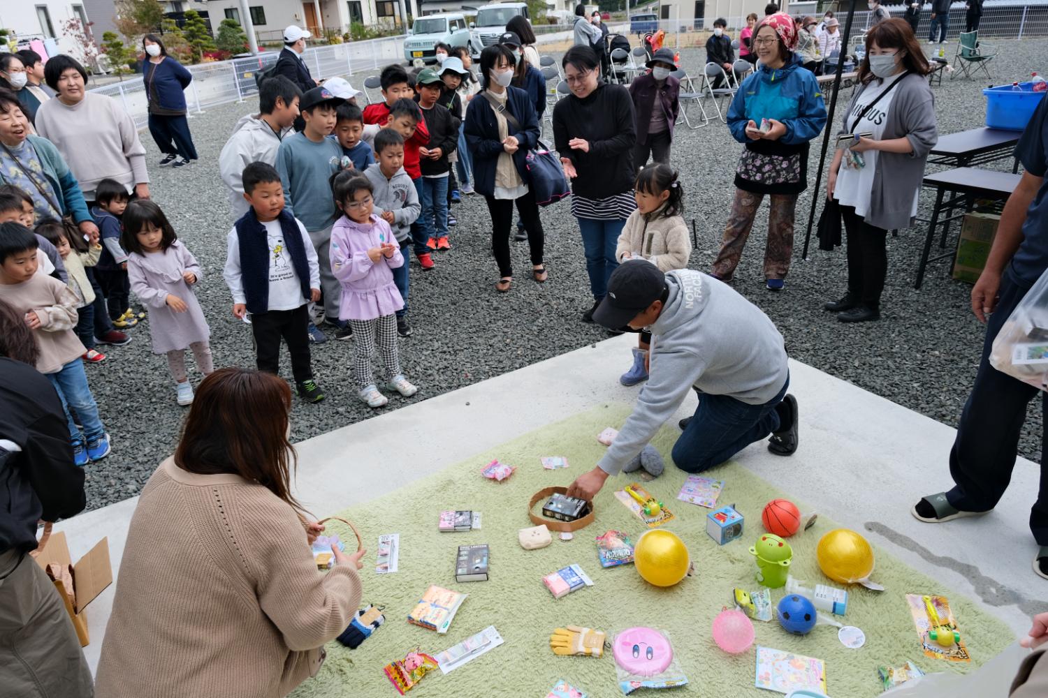 広場で子どもや大人の参加者が輪になっておもちゃや景品を囲み、スタッフがそれらを配っているイベントの写真