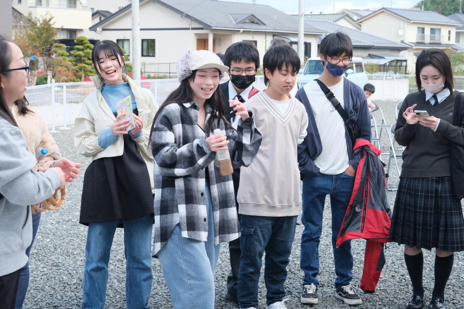 屋外で高校生たちのグループが楽しそうに会話しながら飲み物やスマートフォンを手にしている様子の写真