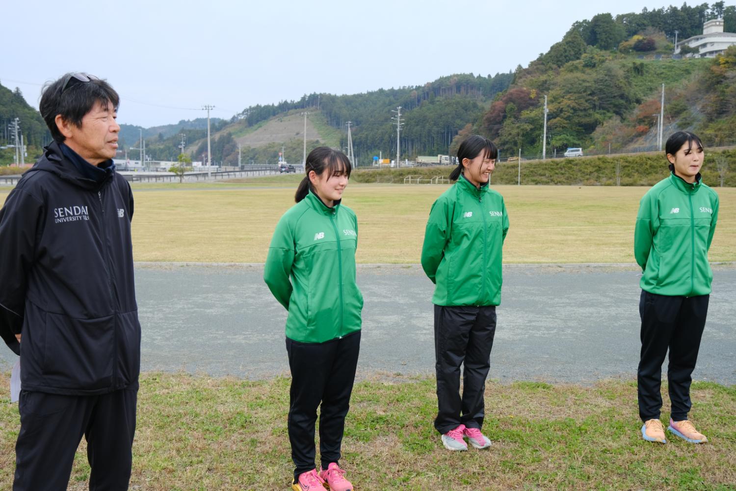 芝生のグラウンドの横に立つ黒と緑のジャージを着た4人が、手を後ろに組んで前を見つめている写真