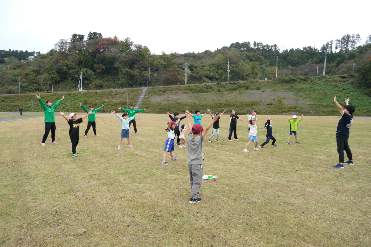 広い芝生の中で、緑のジャージを着た人と子供たちが円を作って体操をしている写真