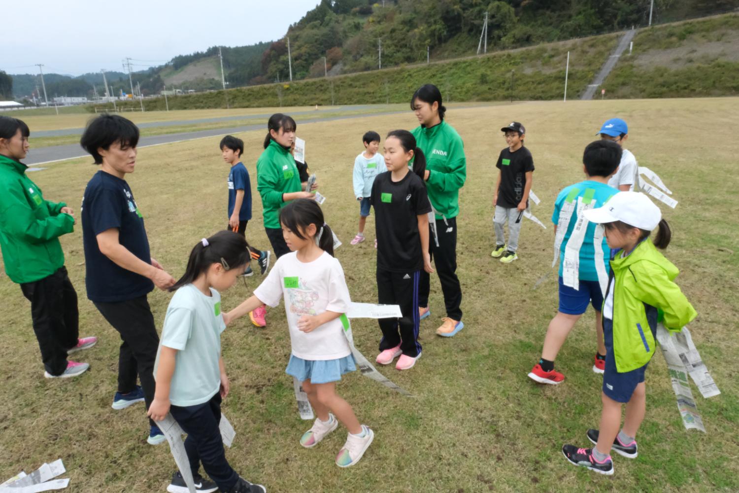 広い芝生の運動場で、大人と子どもたちが新聞紙のしっぽを使った遊びを楽しんでおり、子どもたちはカラフルな運動着を着て笑顔で動き回り、大人たちは緑や黒のジャンパーを着て見守っている写真