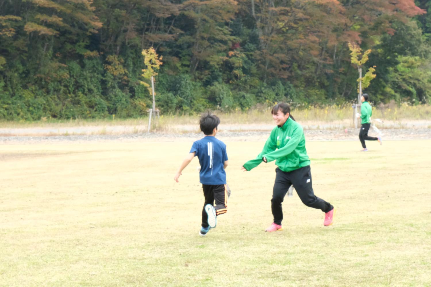 芝生の広場で緑のジャンパーを着た大人が笑顔で男の子を追いかけ、遠くには別の大人が走っている様子が見える写真