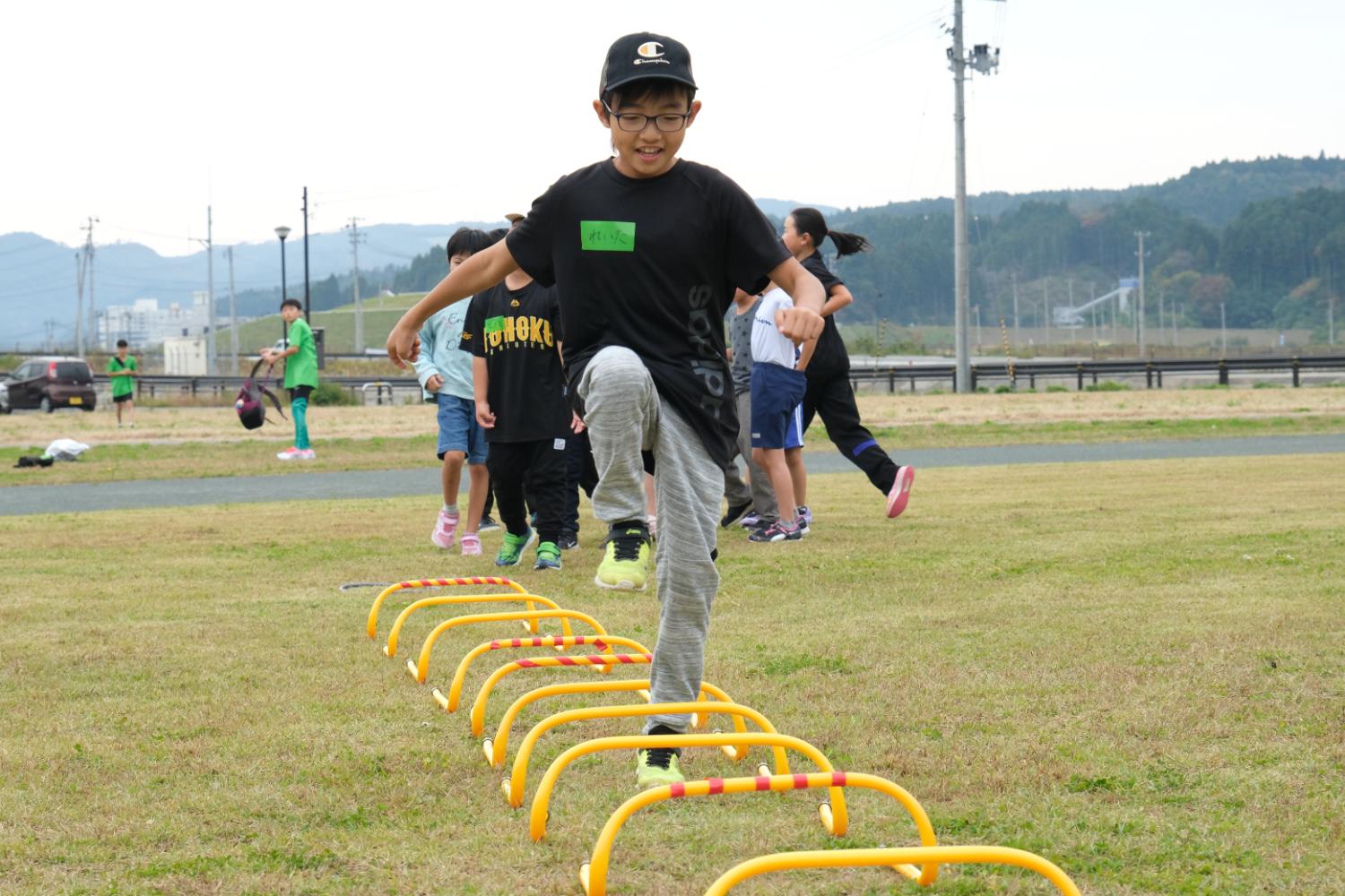 広い芝生の運動場で、男の子が黄色いミニハードルを元気よく跳び越えており、後ろには順番を待つ子どもたちが見える写真