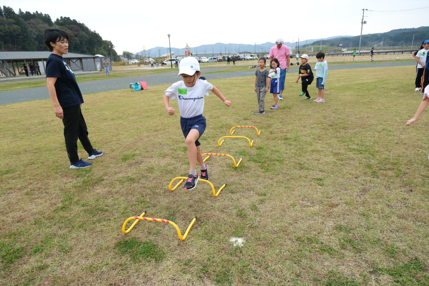 子どもたちが芝生広場で黄色いハードルを順にジャンプして運動している様子で、後ろには見守る大人や待っている子どもたちが並んでいる写真