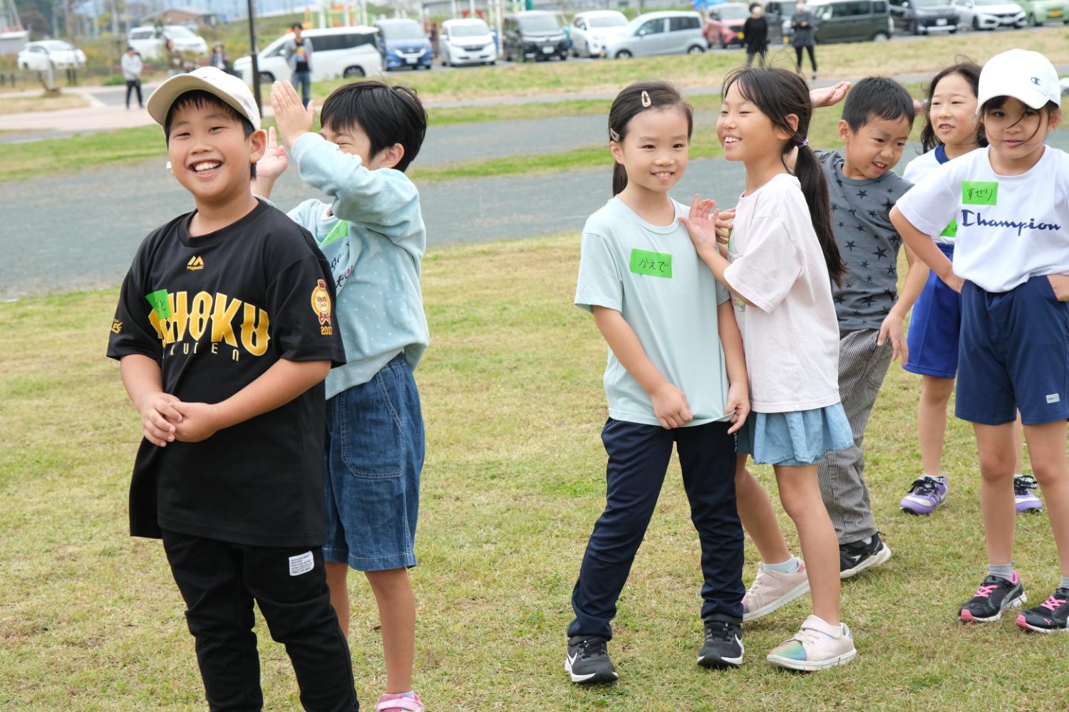自分の名前が書かれたシールを貼った数人の子供たちが屋外の芝生の上に並んでいる写真