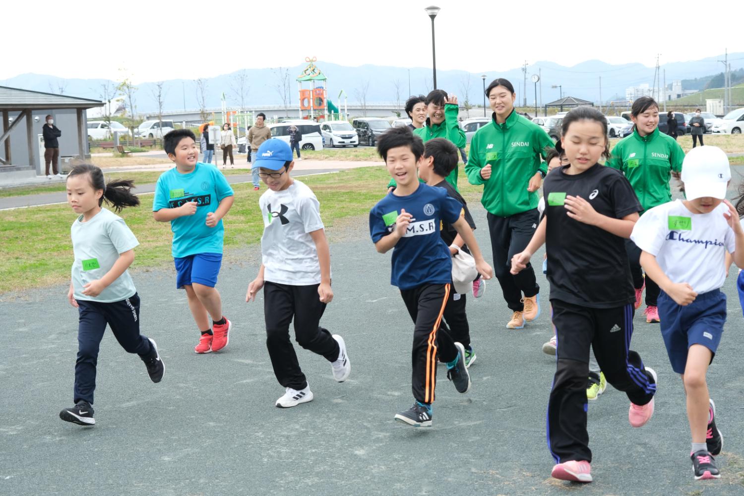 数人の子供たちが屋外の広場で元気に走っていて、その後ろを緑のジャージを着た大人が走っている写真