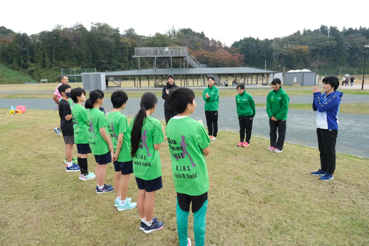 Tシャツを着た中学生の男女と緑のジャージを着た指導者が対面して芝生の運動場に並び、話しながら拍手している写真