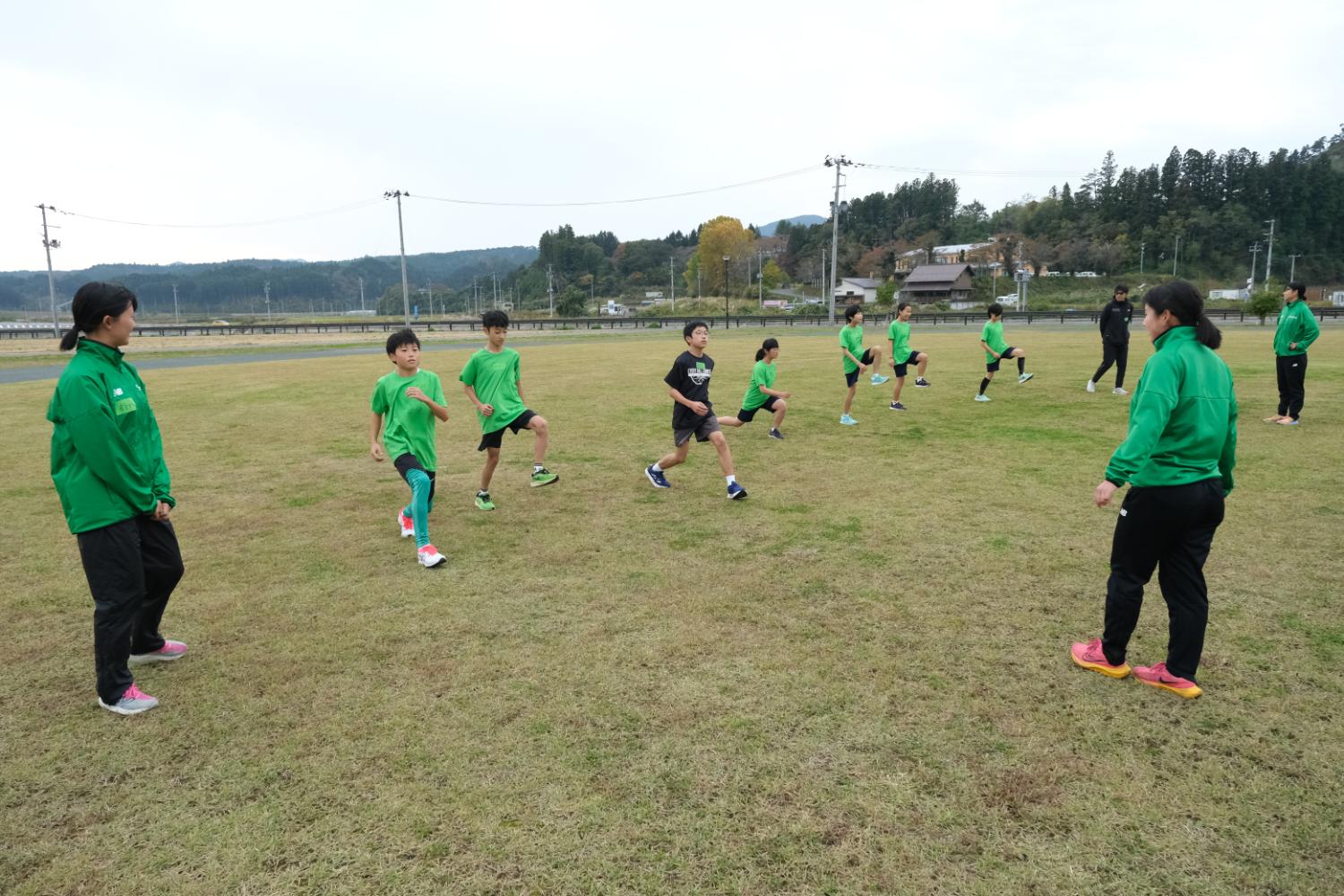 中学生の男女が芝生の運動場に広がって並び、片足を上げてランニングの準備運動をしており、緑色のジャージを着た指導者が両端で見守っている写真