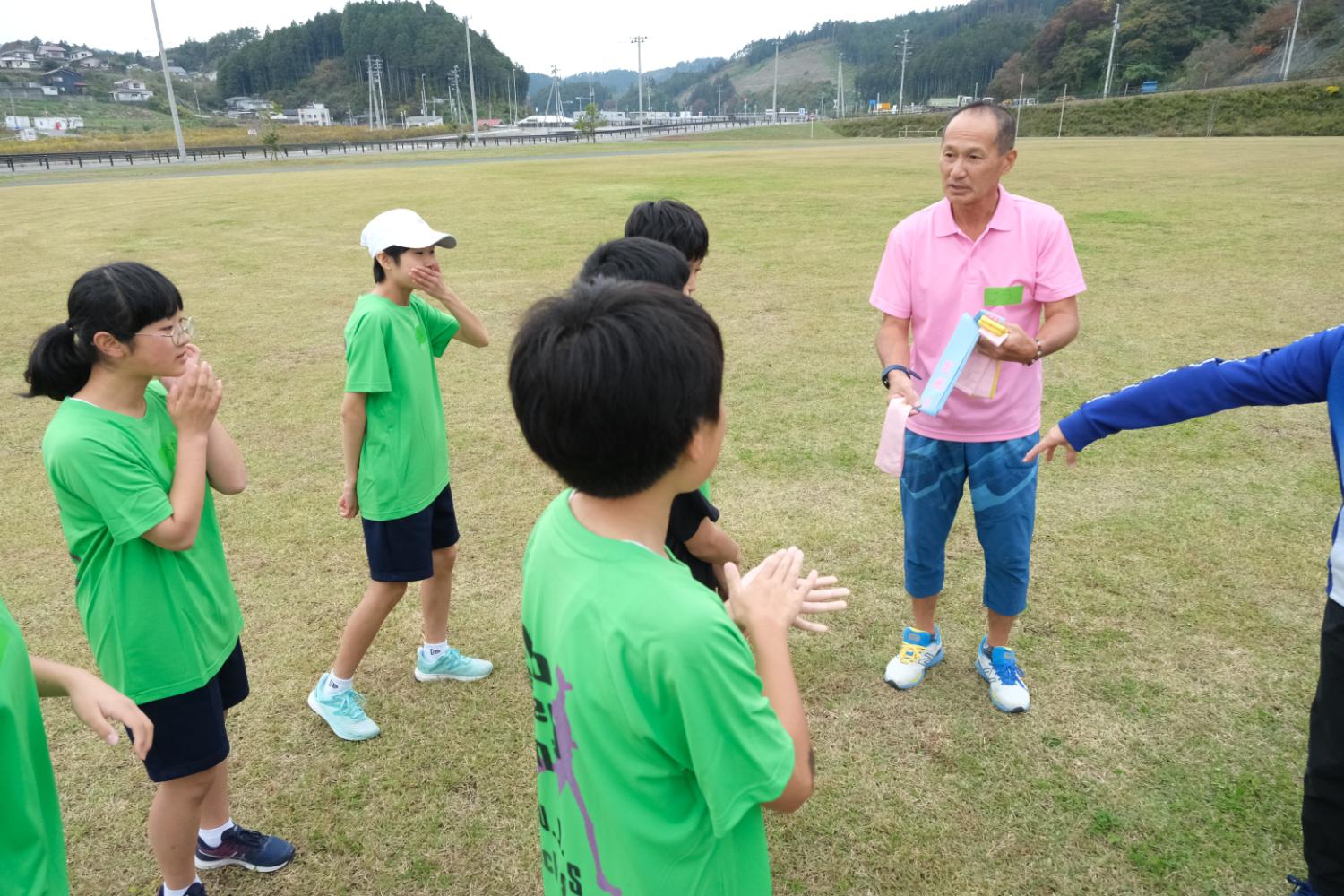 緑のTシャツを着た子どもたちが芝生の広場で、ピンクのポロシャツと青いハーフパンツの指導者から襷を見せてもらい準備をしている写真
