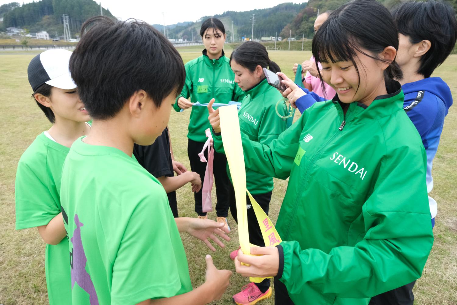 緑色のジャージを着た若い女性が黄色い襷を子どもに渡しており、周囲には同じ服装の仲間と青い服の人が立っている写真