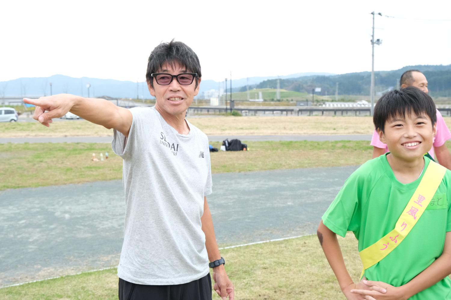 男性が指を指して隣に立っている緑色のTシャツを着て黄色の襷をかけた子供に何かを説明している写真