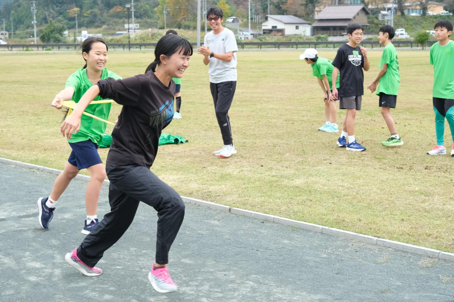 広い芝生のフィールドで、緑のシャツを着た子どもたちや大人の男性が見守る中、緑のTシャツの着た女の子から襷をもらった黒いTシャツの女の子が走り出す写真