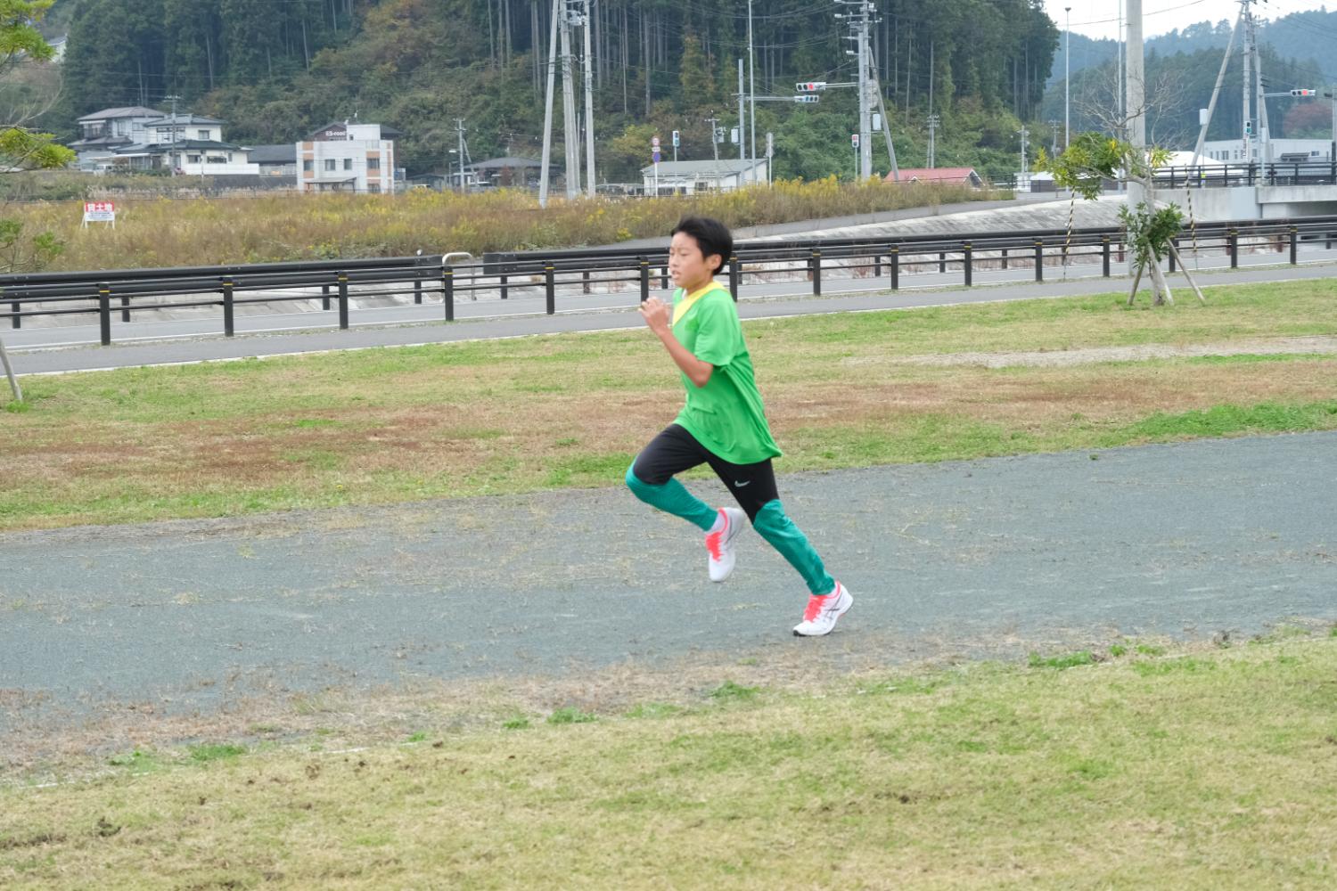 陸上トラックで、緑のシャツとタイツを着て黄色の襷をかけた男の子が真剣な表情で全力で走っている写真