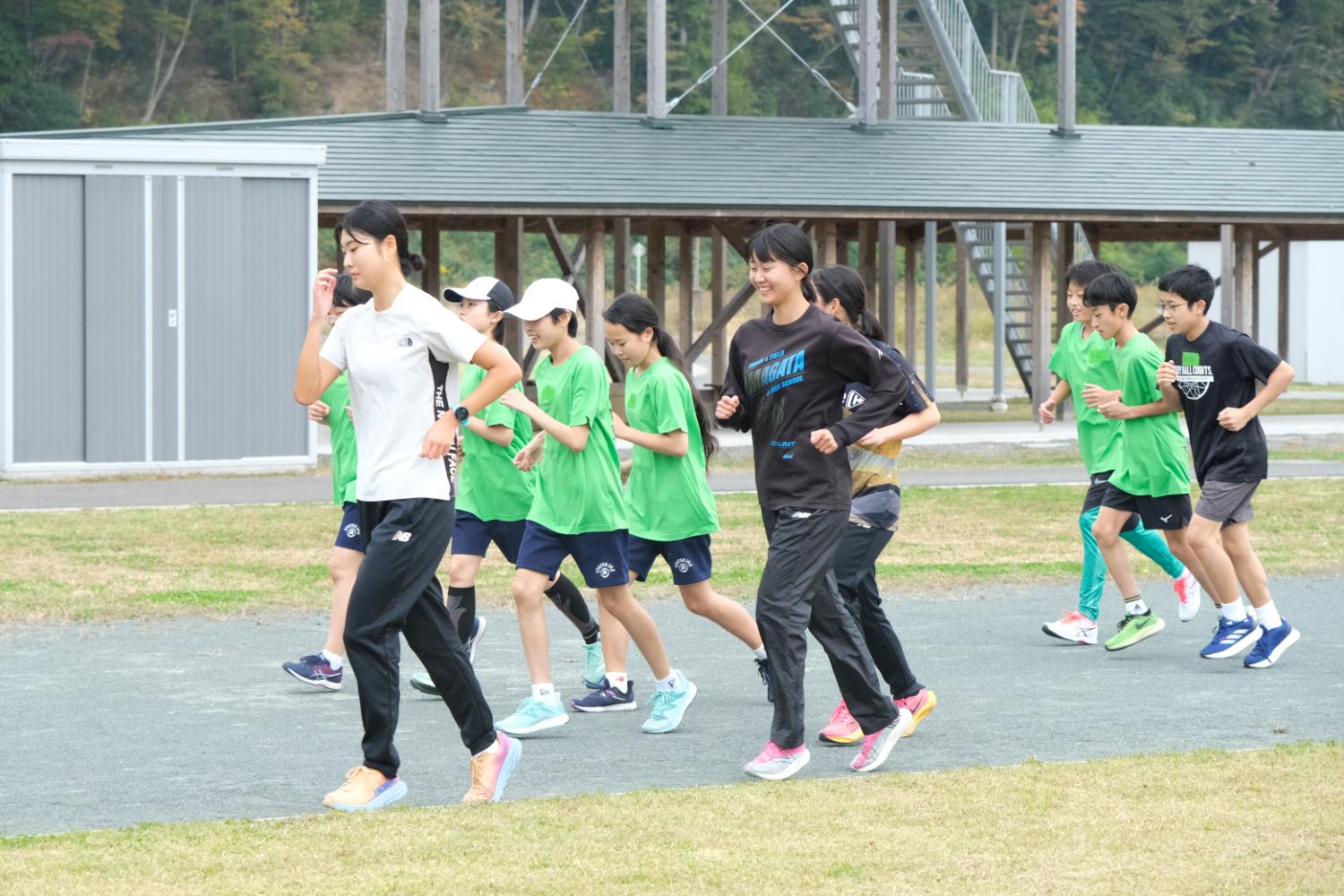 トラックで白や黒、緑のシャツを着た子どもたちが一列になって楽しそうにランニングしている秋の屋外トレーニング風景の写真