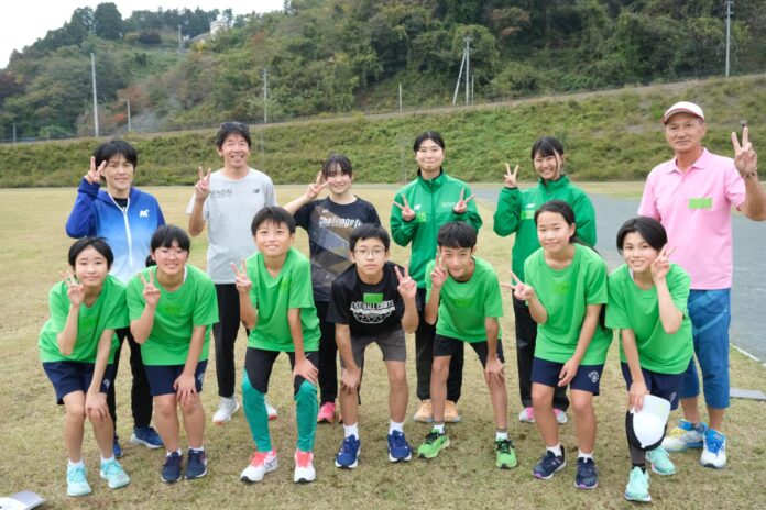 芝生の広場に立つ13人の男女が笑顔でピースサインをしており、前列には緑の運動着を着た子どもたちがしゃがみ、後列には指導者と思われる大人や女子生徒が立っている写真