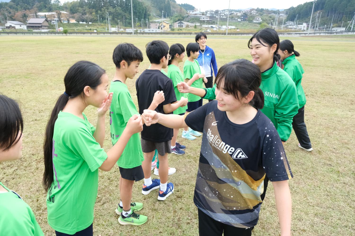 広い芝生のグラウンドで、緑や黒のTシャツを着た子どもたちと緑のジャージを着た指導者が向かい合ってグータッチをしながら交流している写真