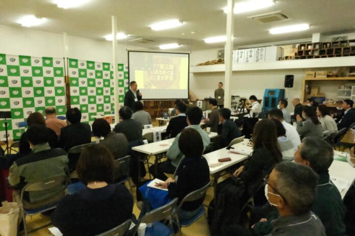 地域住民が参加する講演会で、講師がスクリーンを使って『第三回いのちをつなぐまち学会』について話している様子を、正面から撮影した室内の写真