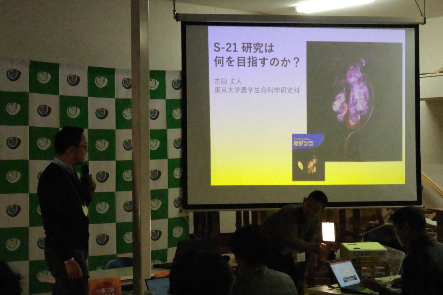 マイクを持った発表者が『S-21研究は何を目指すのか？』というタイトルのスライドを指しながら説明しており、背景には自治体のロゴが入った緑と白の幕が張られている発表の写真