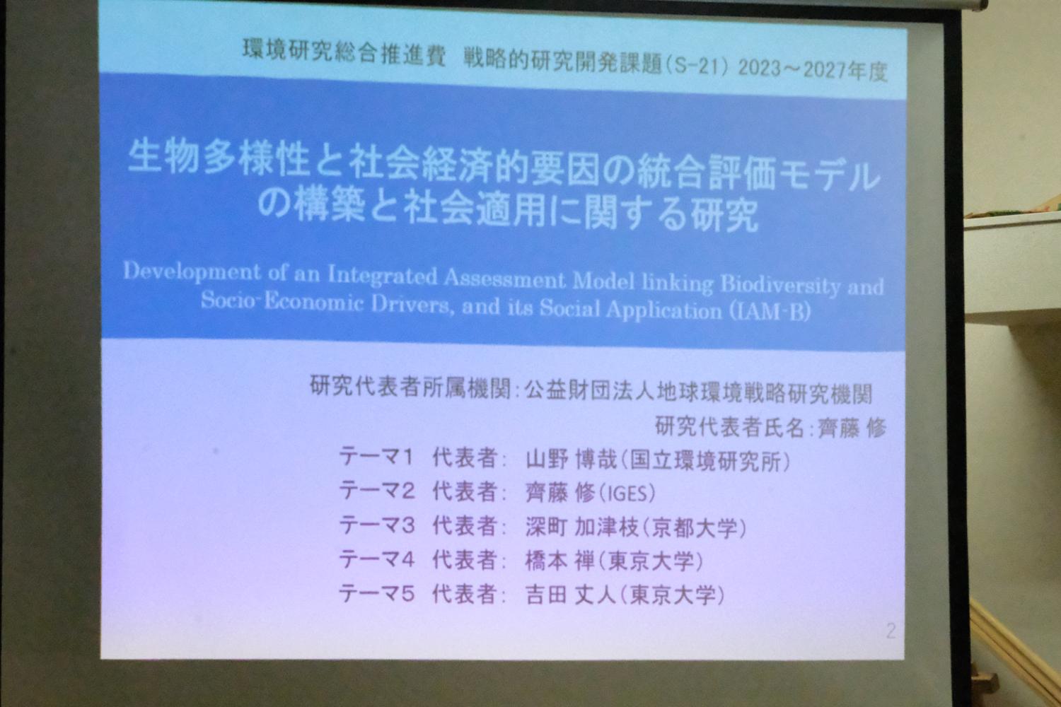 生物多様性と社会経済的要因の統合評価モデルの構築と社会適用に関する研究』という研究課題タイトルがスライドに大きく表示されており、代表者名や所属機関などの詳細が並んでいる研究発表のスライドの写真