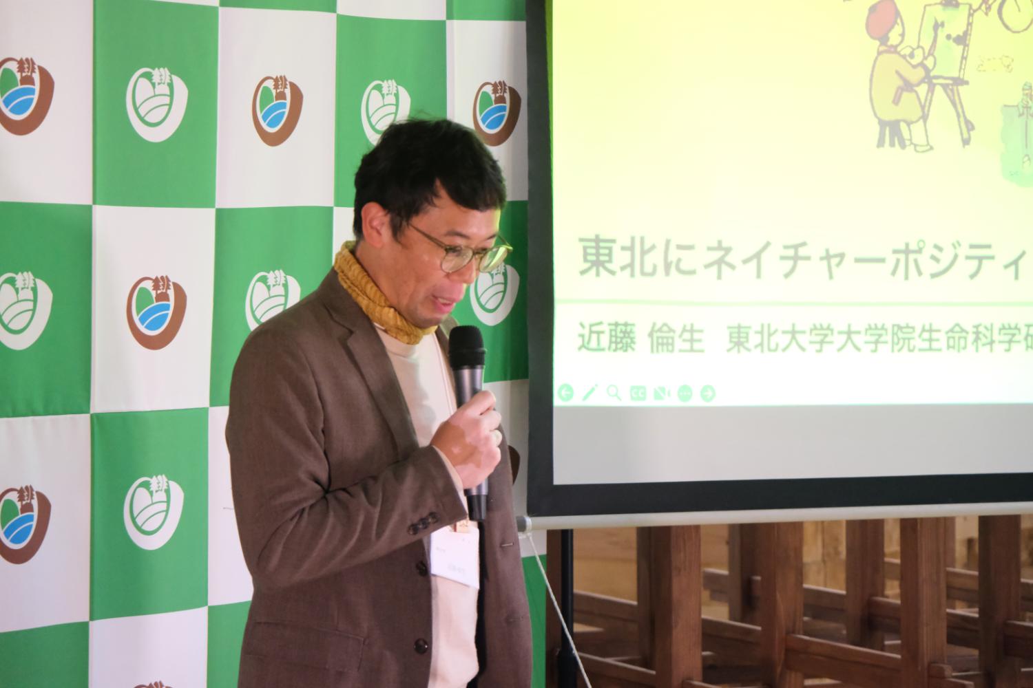 東北にネイチャーポジティブを』というスライドを背景に、マイクを持って発表している男性講演者の写真