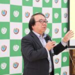 宮城大学のロゴが入った緑と白のチェック柄のパネルの前に黒いスーツを着た男性が右手にマイクを持ち、右手を上げながら話をしている写真（DSCF9435のページへリンク）