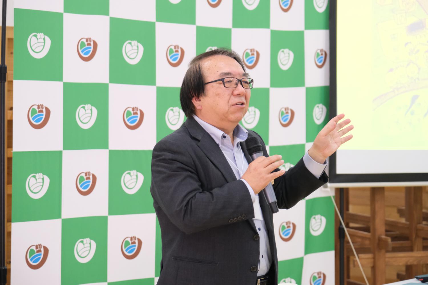 宮城大学のロゴが入った緑と白のチェック柄のパネルの前に黒いスーツを着た男性が右手にマイクを持ち、右手を上げながら話をしている写真