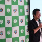 宮城大学のロゴが入った緑と白のチェック柄のパネルの前に黒いジャケットと白いシャツを着た男性がマイクを手に笑顔で話している写真（DSCF9443のページへリンク）