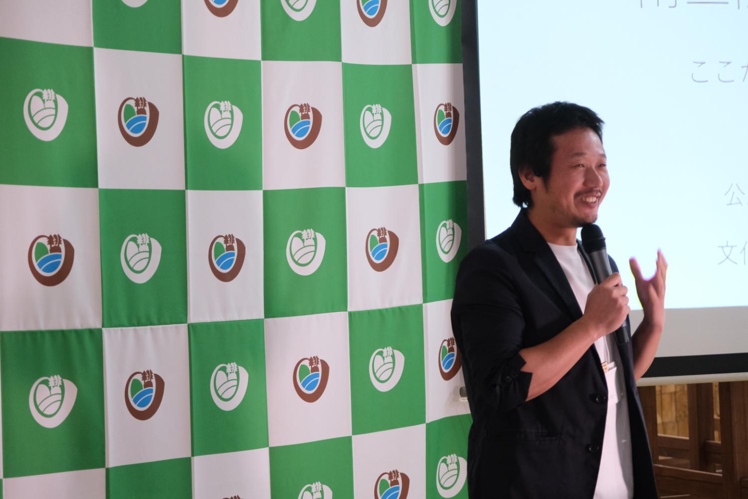 宮城大学のロゴが入った緑と白のチェック柄のパネルの前に黒いジャケットと白いシャツを着た男性がマイクを手に笑顔で話している写真