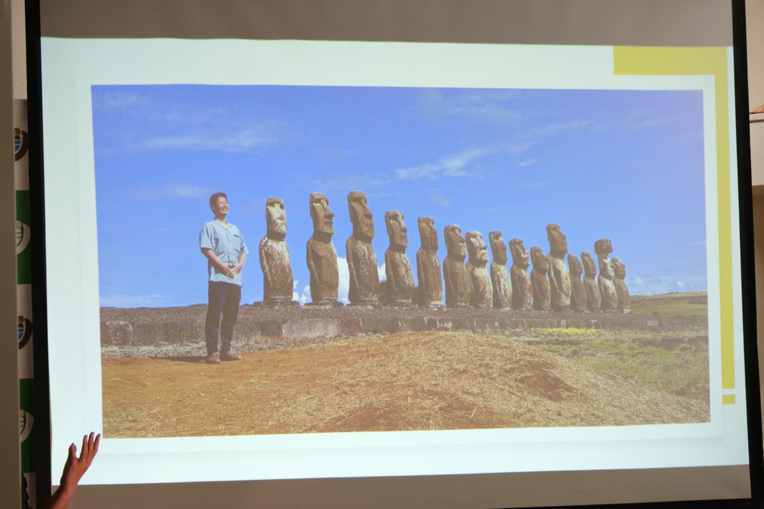 青空の下で乾いた赤土の大地で一列に並んだ15体のモアイ像と一緒に左側に男性が同じ方向を見て立っている写真