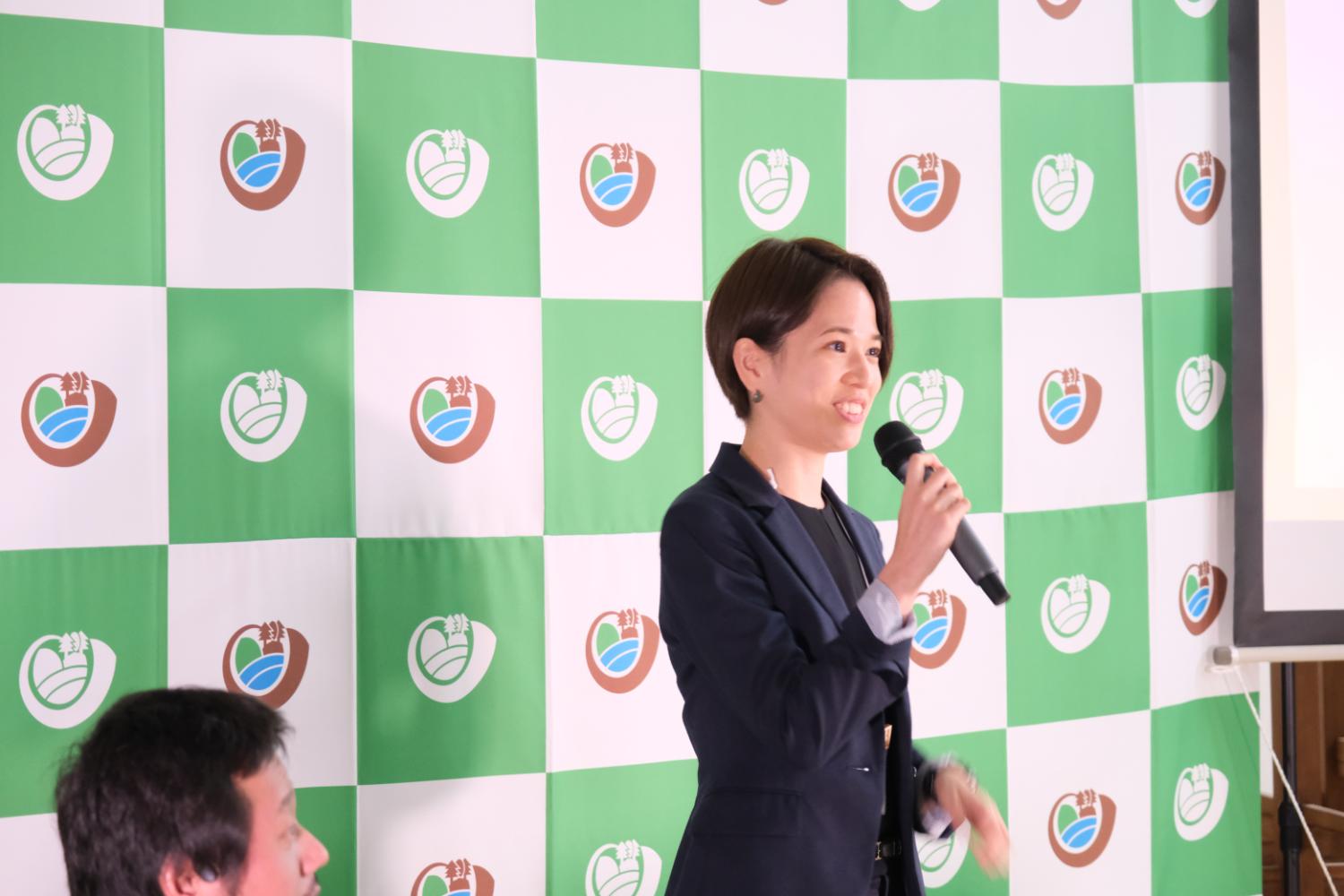宮城大学のロゴが入った緑と白のチェック柄のパネルの前に黒いスーツを着た女性が右手にマイクを持ち、話をしている写真