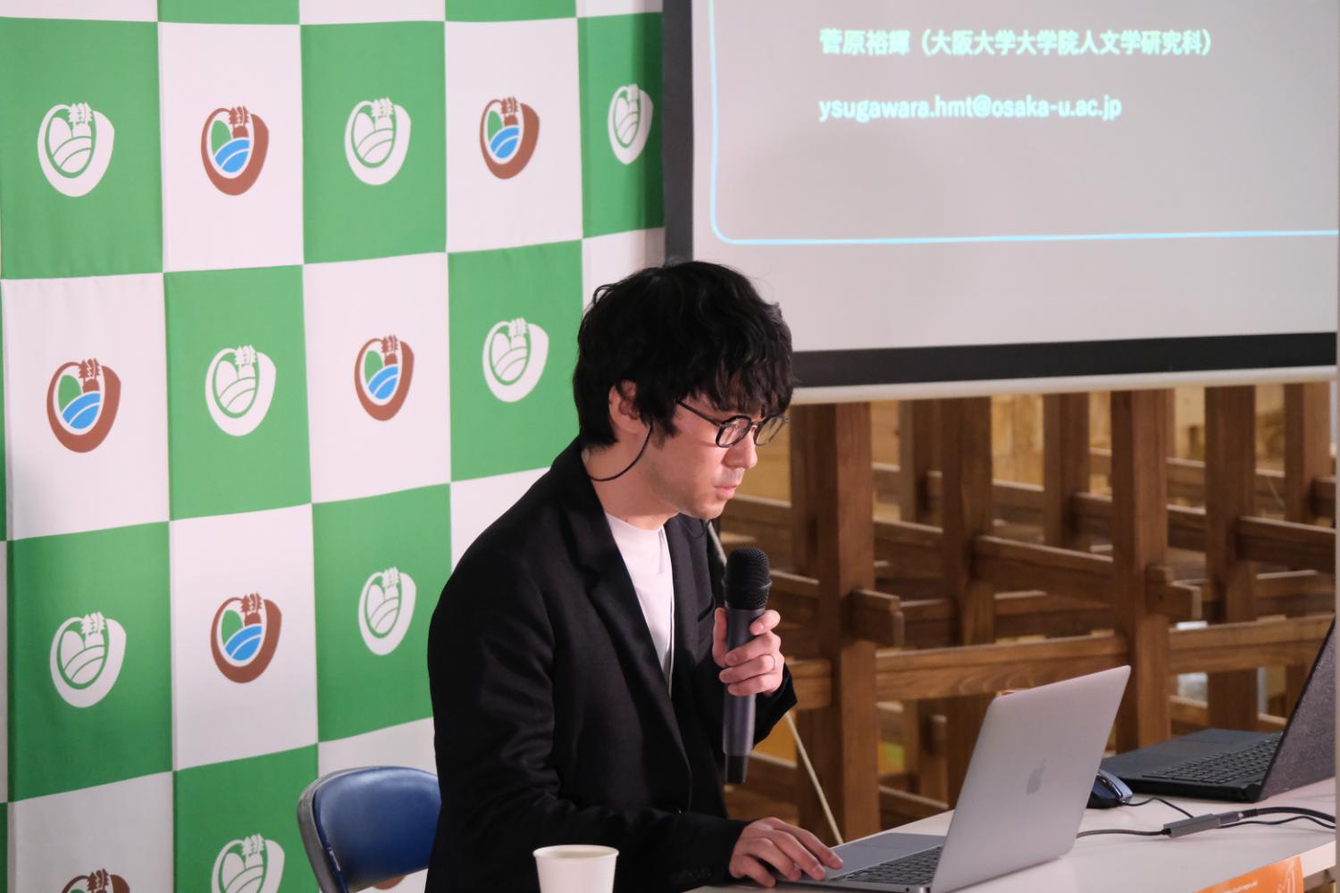 宮城大学のロゴが入った緑と白のチェック柄のパネルの前にマイクを持った男性がノートパソコンを操作しながら話している写真