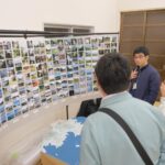 数人の男女が室内で地図の模型の前に立ち、壁に並べられた多くの風景写真を見て話している様子の写真（DSCF9517のページへリンク）