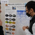 ボードに貼られた「環境DNA調査で海の変化をモニタリング」と書かれ複数の魚の写真が掲載された掲示物を2人の男性が見ている写真（DSCF9535のページへリンク）