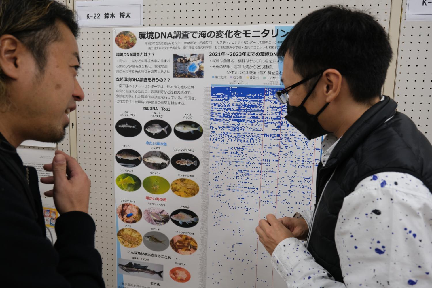 ボードに貼られた「環境DNA調査で海の変化をモニタリング」と書かれ複数の魚の写真が掲載された掲示物を2人の男性が見ている写真