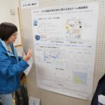 ボードに貼られた「カキの養殖の意思決定に関する進化ゲーム理論解析」と書かれた掲示物を女性が指しながら説明をしているのを2人の方が聞いている写真（DSCF9539のページへリンク）