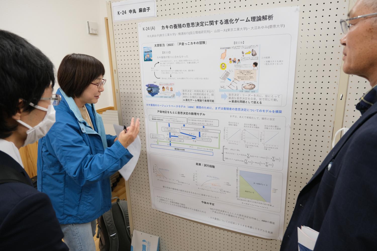 ボードに貼られた「カキの養殖の意思決定に関する進化ゲーム理論解析」と書かれた掲示物を女性が指しながら説明をしているのを2人の方が聞いている写真