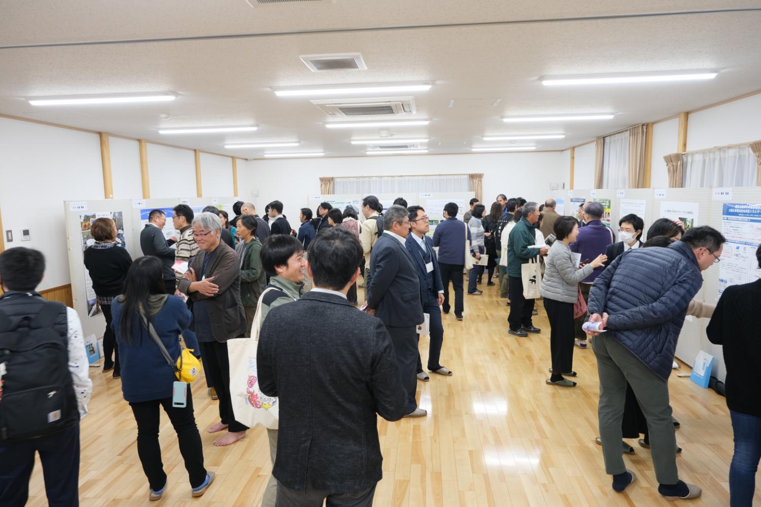 室内の壁際全体にボードが置かれ多くの人々がボードに貼られた掲示物を見ている写真