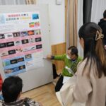 緑色の服を着た人が展示パネルの前でしゃがみながら説明をしており、数人の来場者がその話に耳を傾けている様子の写真（DSCF9569のページへリンク）