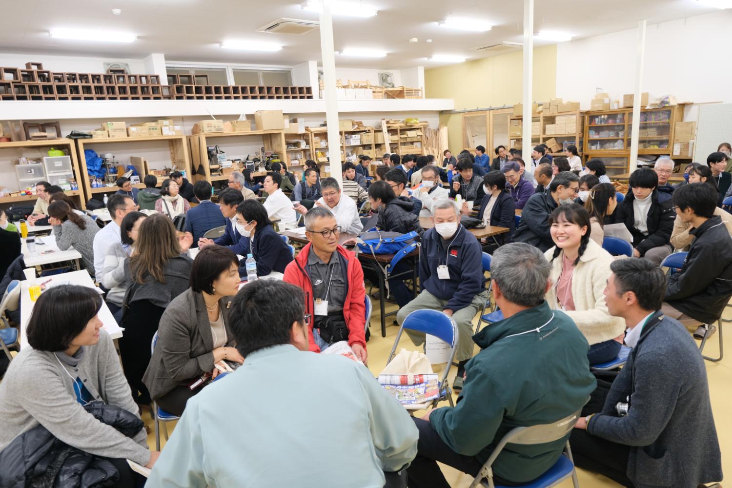 多数の人々が室内の会場で四角形のテーブルを囲んだり、椅子を寄せ円の形にしたグループごとに座って真剣に話し合いや交流をしている様子が写された写真