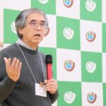 緑と白の市松模様のボードの前で名札を首から下げた男性がマイクを持ち、真剣な表情で話している写真（DSCF9672のページへリンク）