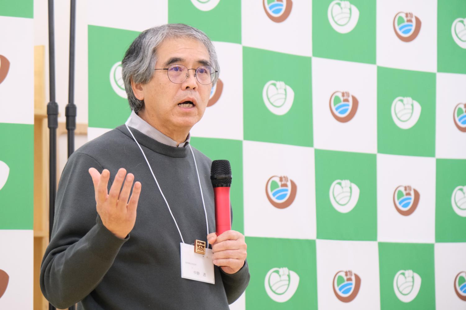 名札を首から下げた男性がマイクを持ち、真剣な表情で話している写真