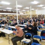 長机と青い椅子が並べられた会場で、多くの人が前方に注目しながら拍手をしている写真（DSCF9674のページへリンク）