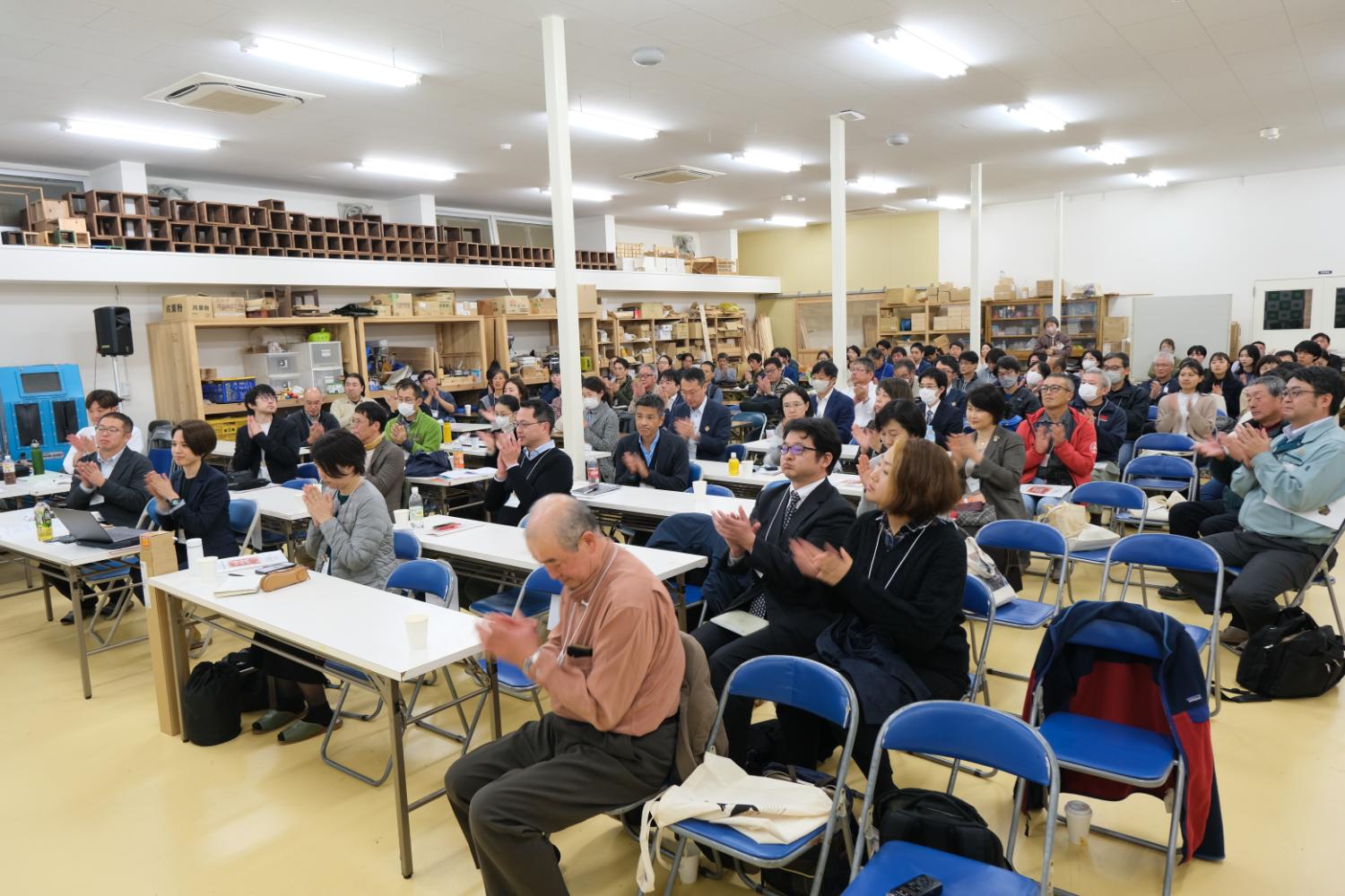長机と青い椅子が並べられた会場で、多くの人が前方に注目しながら拍手をしている写真