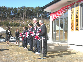 晴天のもと、新しく完成した三陸町図書館の前で関係者の方々が並び、紅白のテープをカットしている落成式の様子を写した写真。
