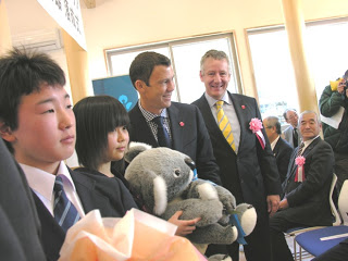 コアラのぬいぐるみを抱える女子学生の後ろに、笑顔で立つブルース・ミラー駐日米大使と関係者男性が写っている落成式の様子の写真