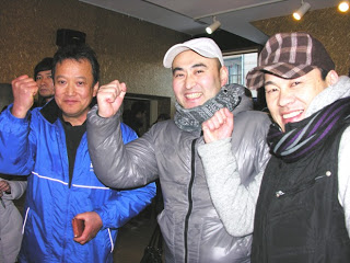 青いジャケット、灰色のジャケットに白い帽子、黒いベストにチェック柄の帽子を身につけた三人の男性が笑顔でガッツポーズをしている写真